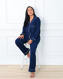 Woman wearing navy blue pajamas with white piping sitting on a stool against a white wall.