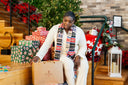 A woman in a white outfit and a colorful knit scarf sits on wooden stairs next to a Christmas tree and wrapped gifts, holding a brown paper gift bag with the 'Heartline Gift Shop' logo.