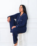 Woman wearing navy blue pajamas with white piping sitting on a white stool against a white wall.